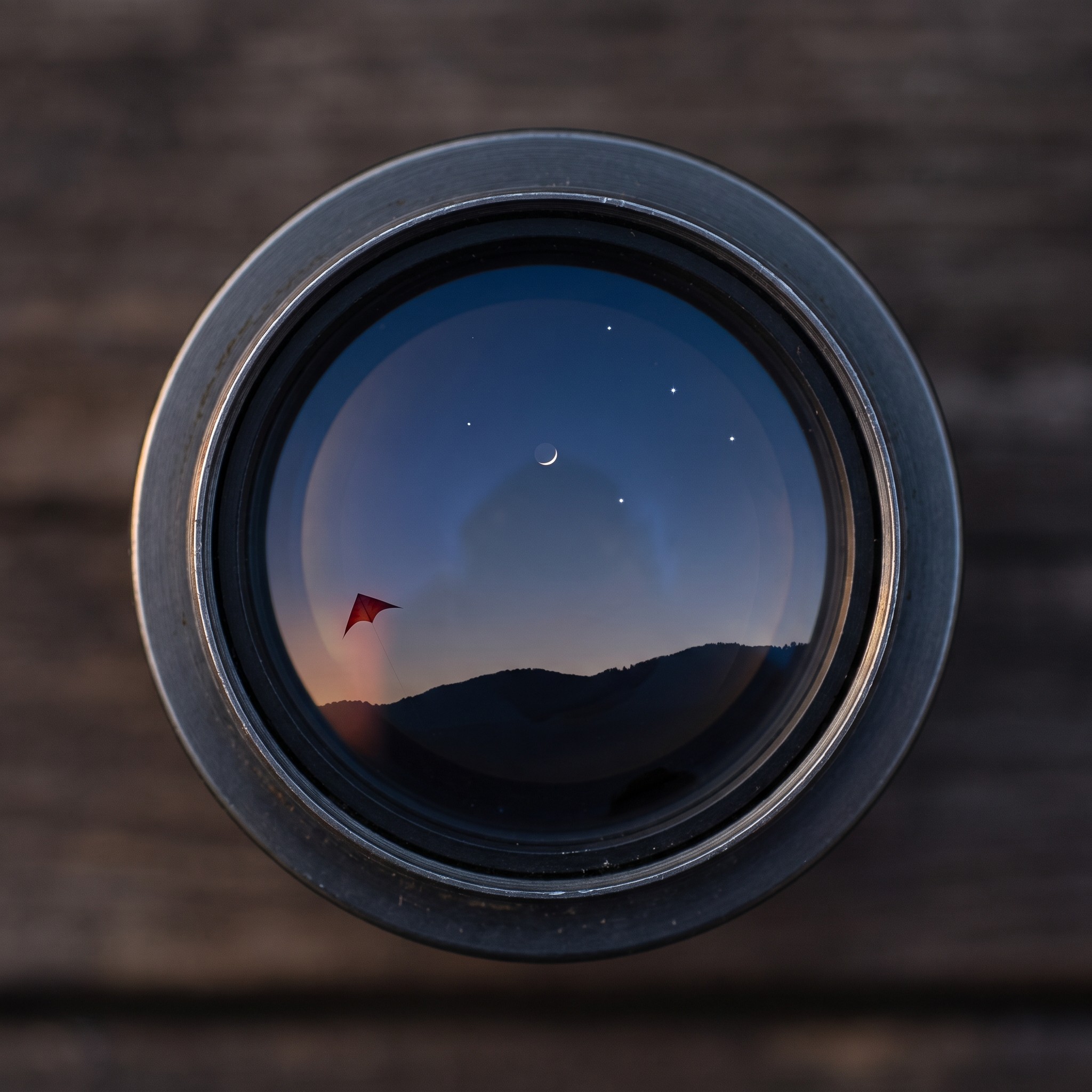 A round eyepiece showing a crescent Moon, a few stars and a red kite over dark hills